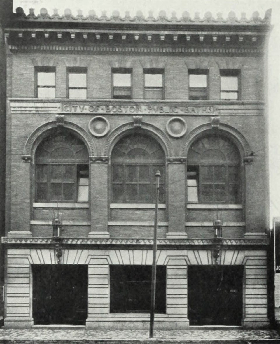 Black-and-white photograph of a three-storey brick and stone civic building seen straight on from the street. Three tall round-arched windows fill the main floor above a rusticated stone ground storey with three dark doorways. A carved stone band above the arches reads “CITY OF BOSTON PUBLIC BATHS”. A street lamp stands on the cobbled pavement in front.